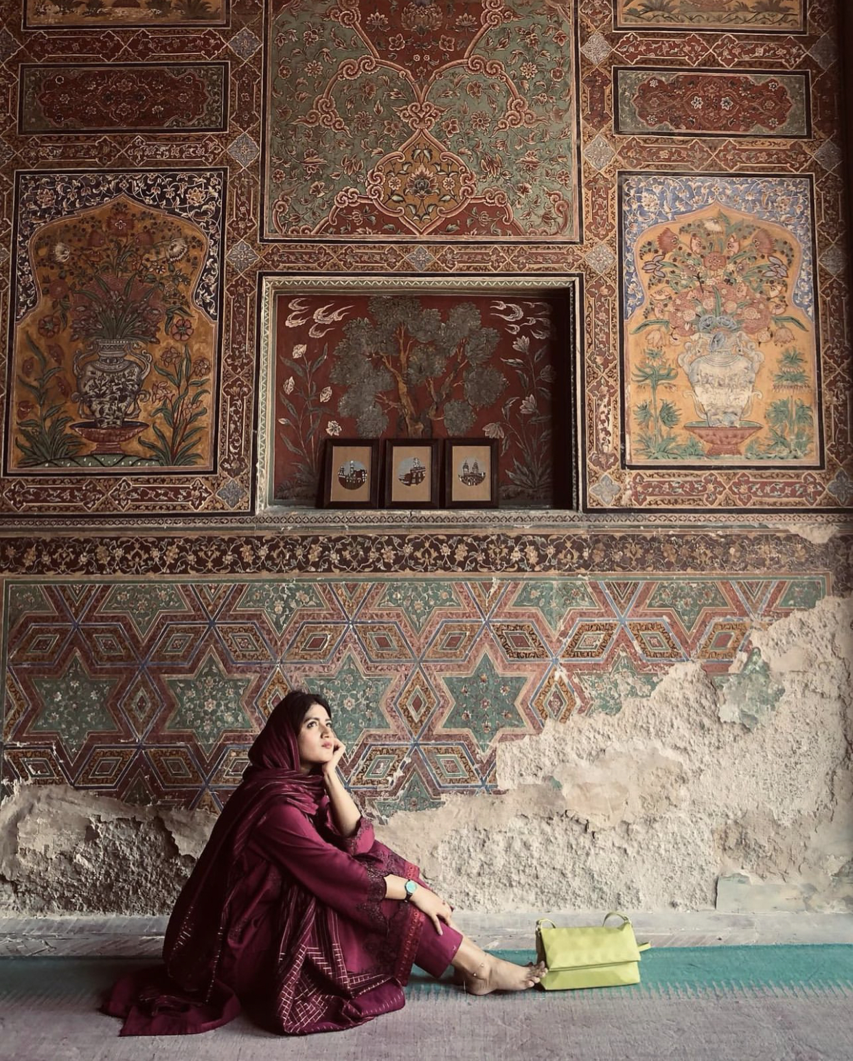 Amna seated in front of hand-painted frescoes with three of her own framed artworks displayed on the shelf behind her